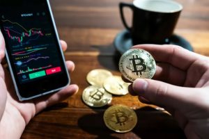 Man holding a bitcoin on one hand and a phone with trading app on the other